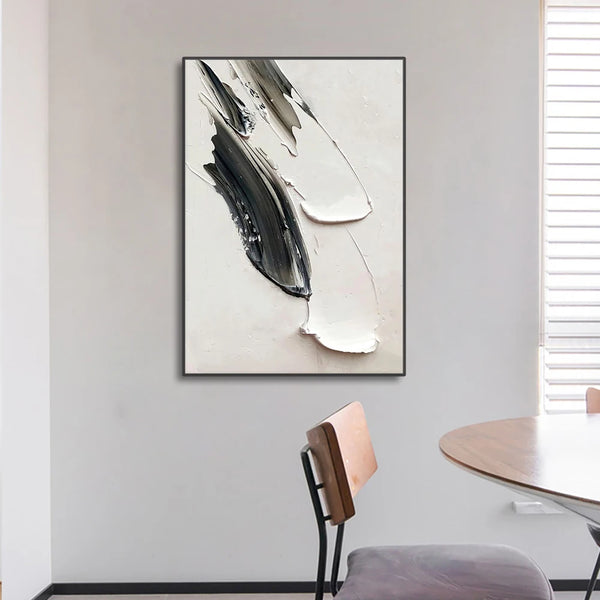 Un tableau de style abstrait noir et blanc est accroché au mur dans une salle à manger. La toile semble avoir reçu de grands coups de pinceaux noirs et blancs ce qui donne un style minimaliste et abstrait à la fois. 
