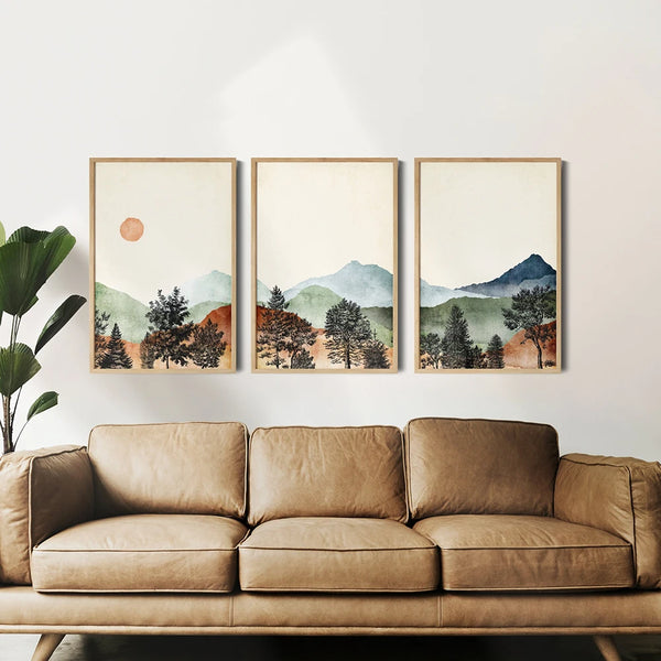 Dans un salon au style bohème trois toiles formant un paysage de montagne aux couleurs automnales et au style abstrait sont affichées au dessus d'un canapé en cuir camel et d'une plante. 