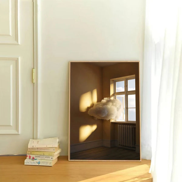 Dans une pièce avec un mur blanc et du parquet, il y a une toile avec un nuage dans un appartement. Il y a à côté des livres. 