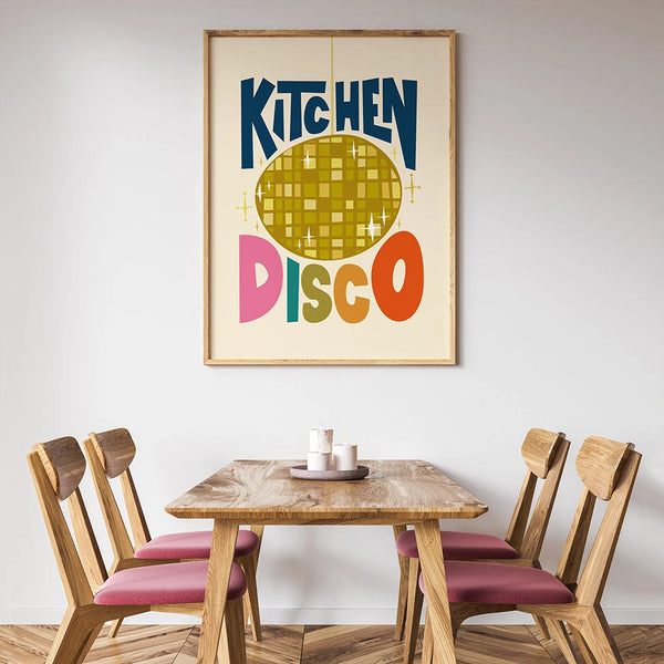 Dans une salle à manger en bois avec des chaises en bois avec des coussins roses, une toile rétro est accrochée. Le tableau représente une boule à facettes notées "Kitchen disco" autour. 