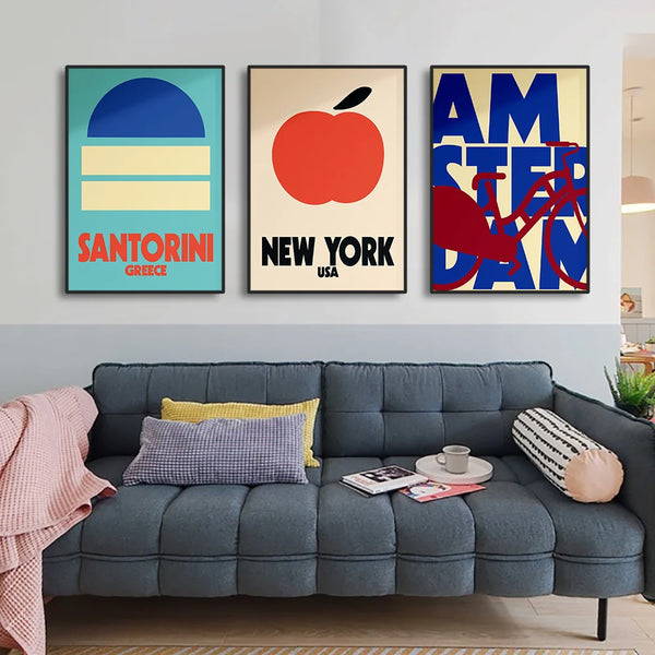 Trois toiles sont accrochées sur un mur au dessus d'un canapé gris. Ce sont trois tableaux représentant chacun une ville dans un style minimaliste et vintage. Il y en a un pour Santorin, un pour New York et un autre pour Amsterdam. 