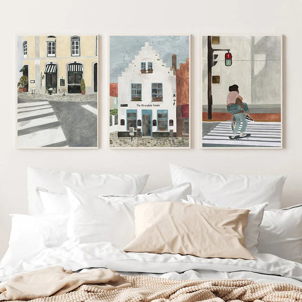 Dans une chambre épurée aux linges de lit blanc et beige, trois toiles sont accrochées au mur. Ce sont des tableaux de style peinture minimaliste représentant des paysages urbains et des couples. 