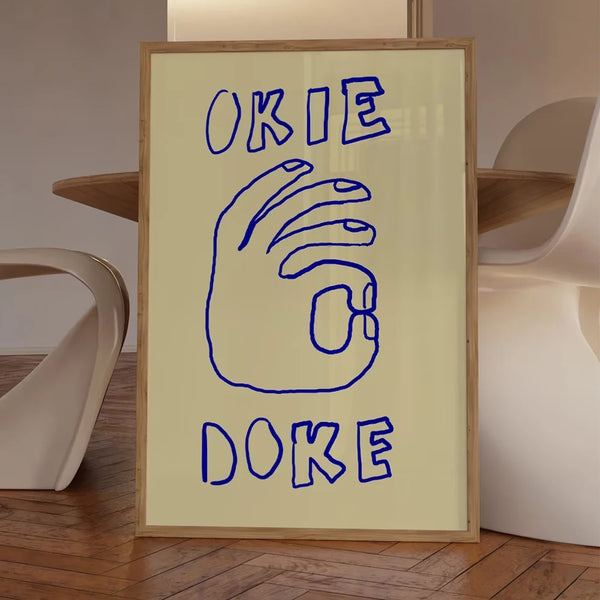 Dans un salon au design scandinave une toile est posé au sol. Il est noté Okie Doke avec une main joignant pouce et index dans un style de dessin minimaliste très tendance. 